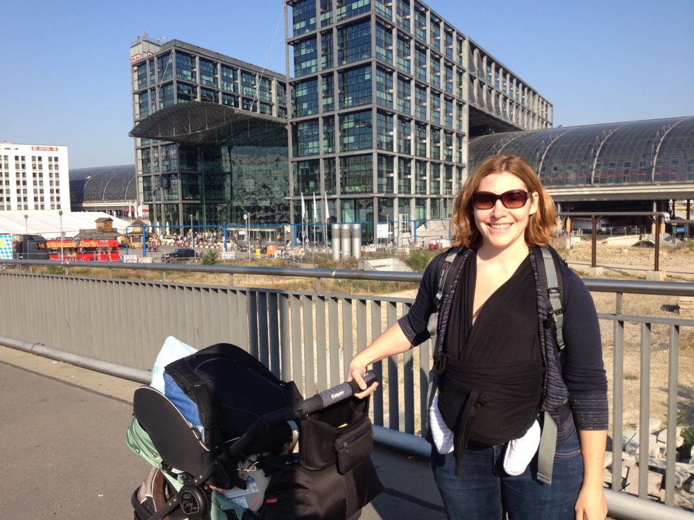 Travel Car Seat Expert - A woman standing on a bridge with a stroller in front of a building, while traveling with her baby.