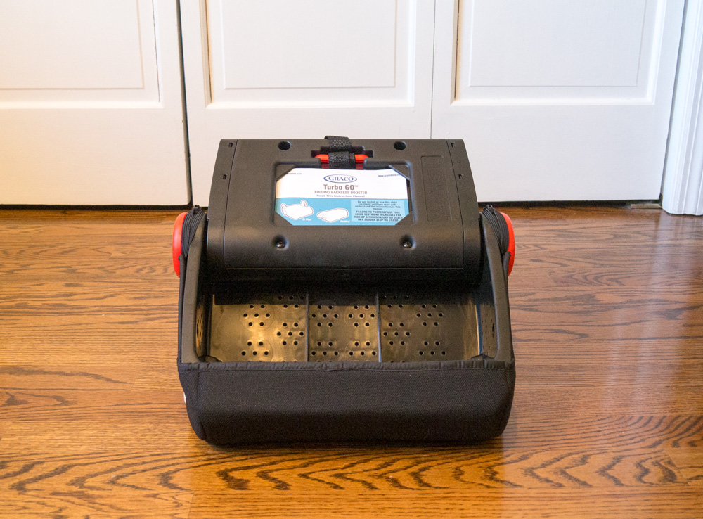 Underside of black Graco Turbo GO backless booster seat: stowed red shoulder belt clip, white and blue manual storage, ventilation holes in underside of booster seat