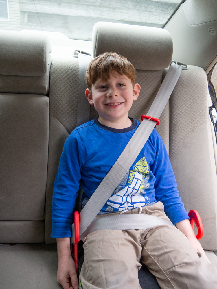 Young boy in blue shirt and tan pants buckled in a tan car, sitting on a Graco Turbo Go backless booster seat with red seatbelt guides visible