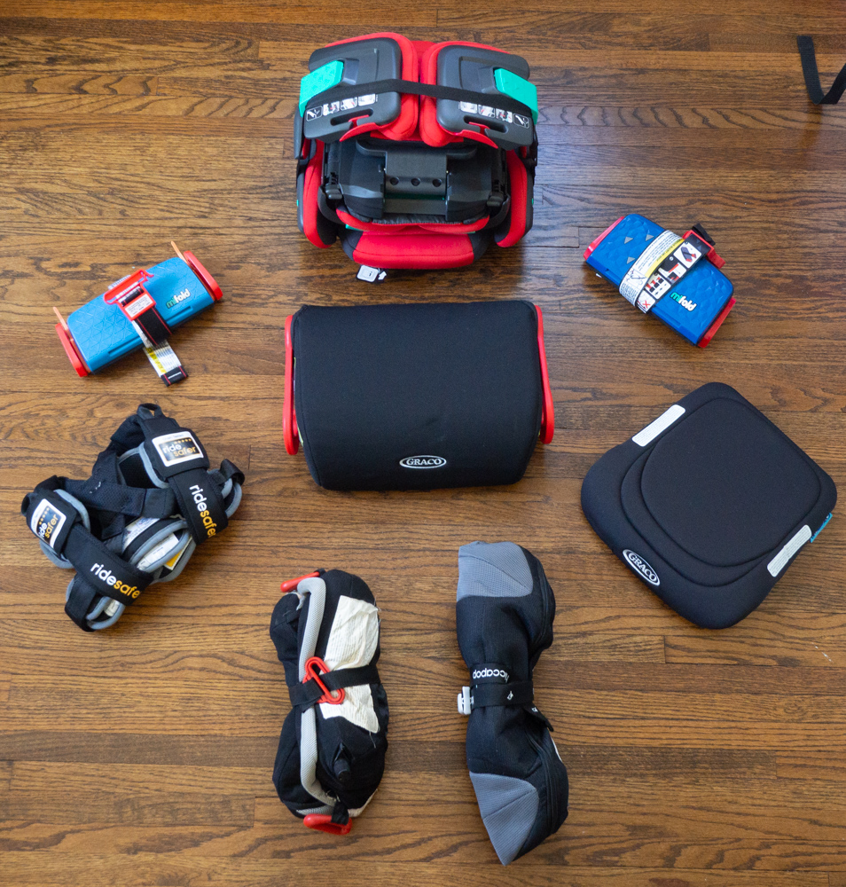 Best travel booster seat options arrayed in a circle on a wood floor, all folded to their most compact sizes. Center: Graco Turbo GO; Clockwise from top: hifold, mifold comfort, Graco RightGuide, hiccapop Uberbooster, Bubblebum, Ride Safer travel vest, mifold original