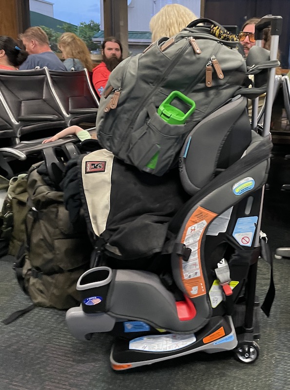 Travel Car Seat Expert - Luggage stacked on a cart at an airport, featuring a large backpack and a child's car seat, along with people sitting in the background. This setup includes 7 genius ideas for how